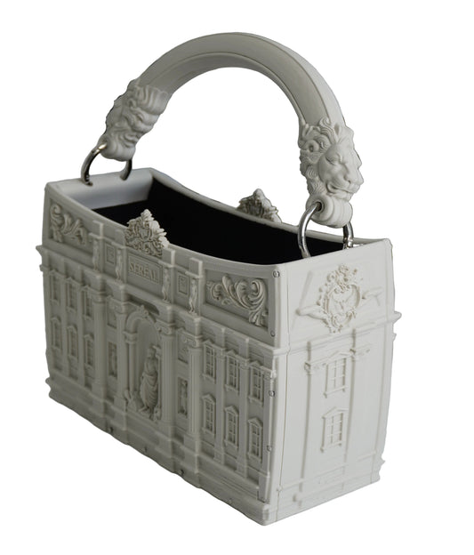 A white decorative rectangular handbag with an ornate classical building facade design and a sculpted top handle, shown on a white surface with green foliage in the background.