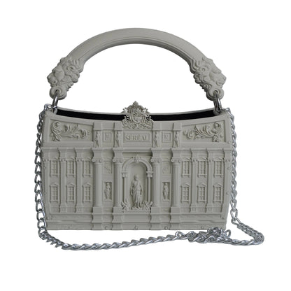 A structured handbag with an intricate architectural building facade design, featuring a curved top handle with sculpted lion details and a silver chain strap, set against a light gray background.