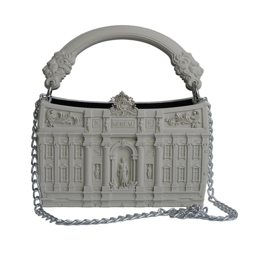 A structured handbag with an intricate architectural building facade design, featuring a curved top handle with sculpted lion details and a silver chain strap, set against a light gray background.