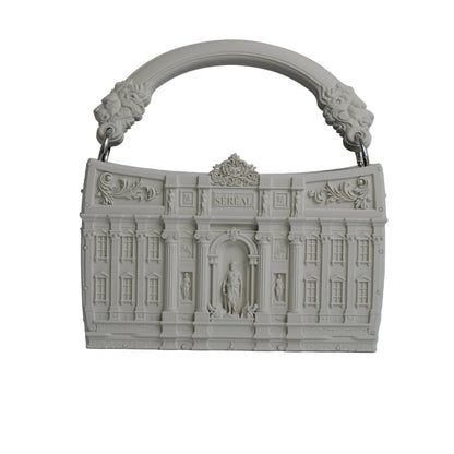 A light gray, sculptural handbag designed to resemble a classical building facade, featuring an integrated arched handle with lion head details, set against a neutral gray background with a soft reflection on the surface below.